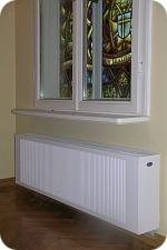 Biały, poziomy grzejnik panelowy zamontowany pod oknem z witrażem w tle, widoczne parkiet i kremowa ściana.