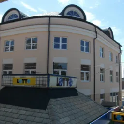 Trzypiętrowy budynek w jasnych kolorach z oknami z białymi ramami, zwieńczony lukarnami, widziany z góry, z billboardem Lotto na pierwszym planie.