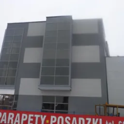 Nowoczesny budynek biurowy z szarą elewacją w geometryczne wzory, widoczny fragment baneru reklamowego u dołu i żółta kabina dźwigu w tle.