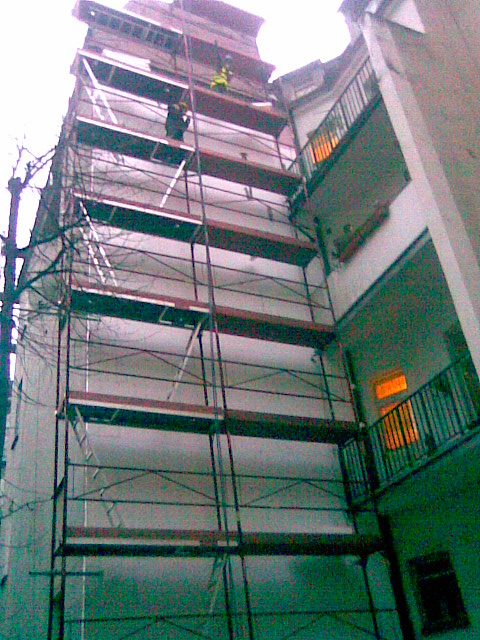 Prace ociepleniowe na elewacji budynku wielorodzinnego z użyciem rusztowania, widoczni dwaj pracownicy w odblaskowych kamizelkach.