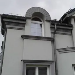 Elewacja budynku z dwoma białymi oknami: dolne prostokątne i g&oacute;rne p&oacute;łokrągłe, ozdobione szarymi detalami architektonicznymi i rynnami, na tle pochmurnego nieba.
