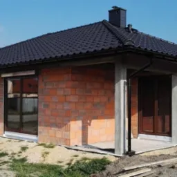 Dom jednorodzinny w trakcie budowy, z zamontowanymi oknami z brązowymi ramami, przesuwne drzwi tarasowe z brązowymi ramami, rolety zwinięte, ściany z czerwonej cegły, czarny dach z kominem.