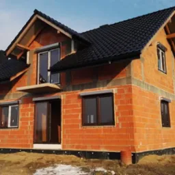 Surowy budynek jednorodzinny w trakcie budowy, z zamontowanymi oknami i roletami zewnętrznymi, widoczne cegły, czarna dachówka i resztki śniegu na ziemi.