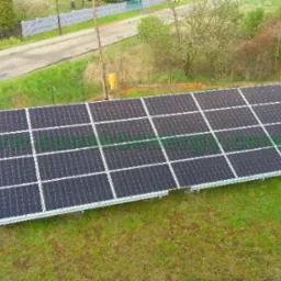 Instalacja paneli słonecznych na trawiastym terenie, widok z góry, w tle dom i ulica.