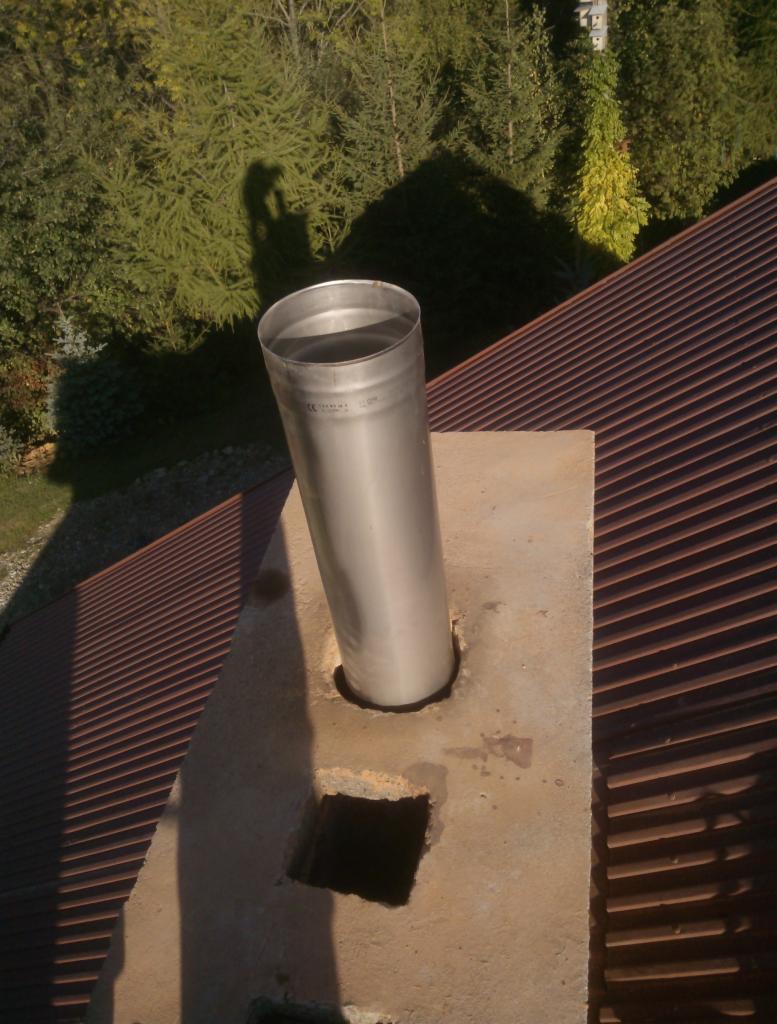 Metalowy wkład kominowy wystający z betonowej obudowy na dachu pokrytym blachodachówką, cień osoby w tle.
