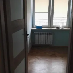 Pokój z parkietem ułożonym w jodełkę, widok zza uchylonych drzwi z szybami, okno z roletami w tle, niebieski parapet z pudełkiem i bibelotami, grzejnik pod oknem, ściany pomalowane na jasny...