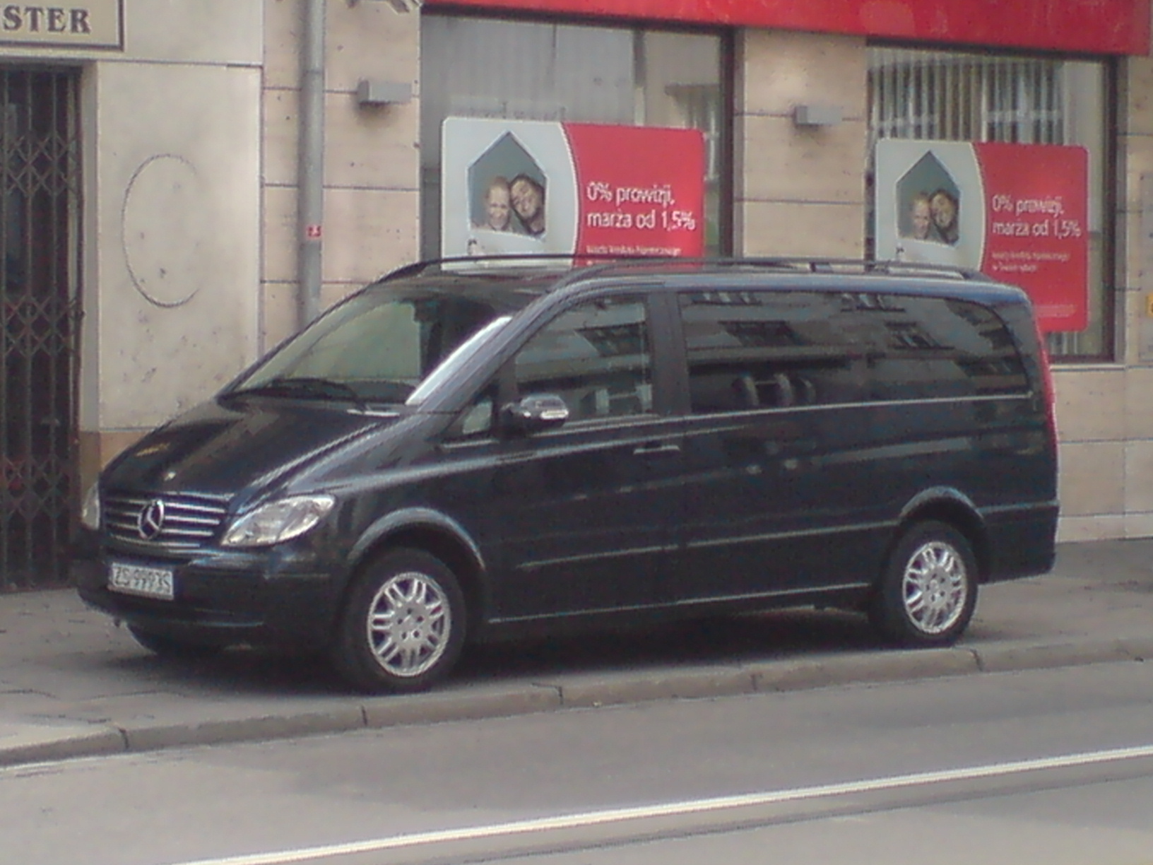 Czarny Mercedes Viano VIP zaparkowany na chodniku przed budynkiem z reklamami bankowymi w tle, widok z boku.