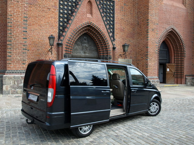 Czarny Mercedes Viano VIP z otwartymi drzwiami przesuwnymi, zaparkowany na brukowanym dziedzińcu przed ceglanym budynkiem z gotyckimi łukami.