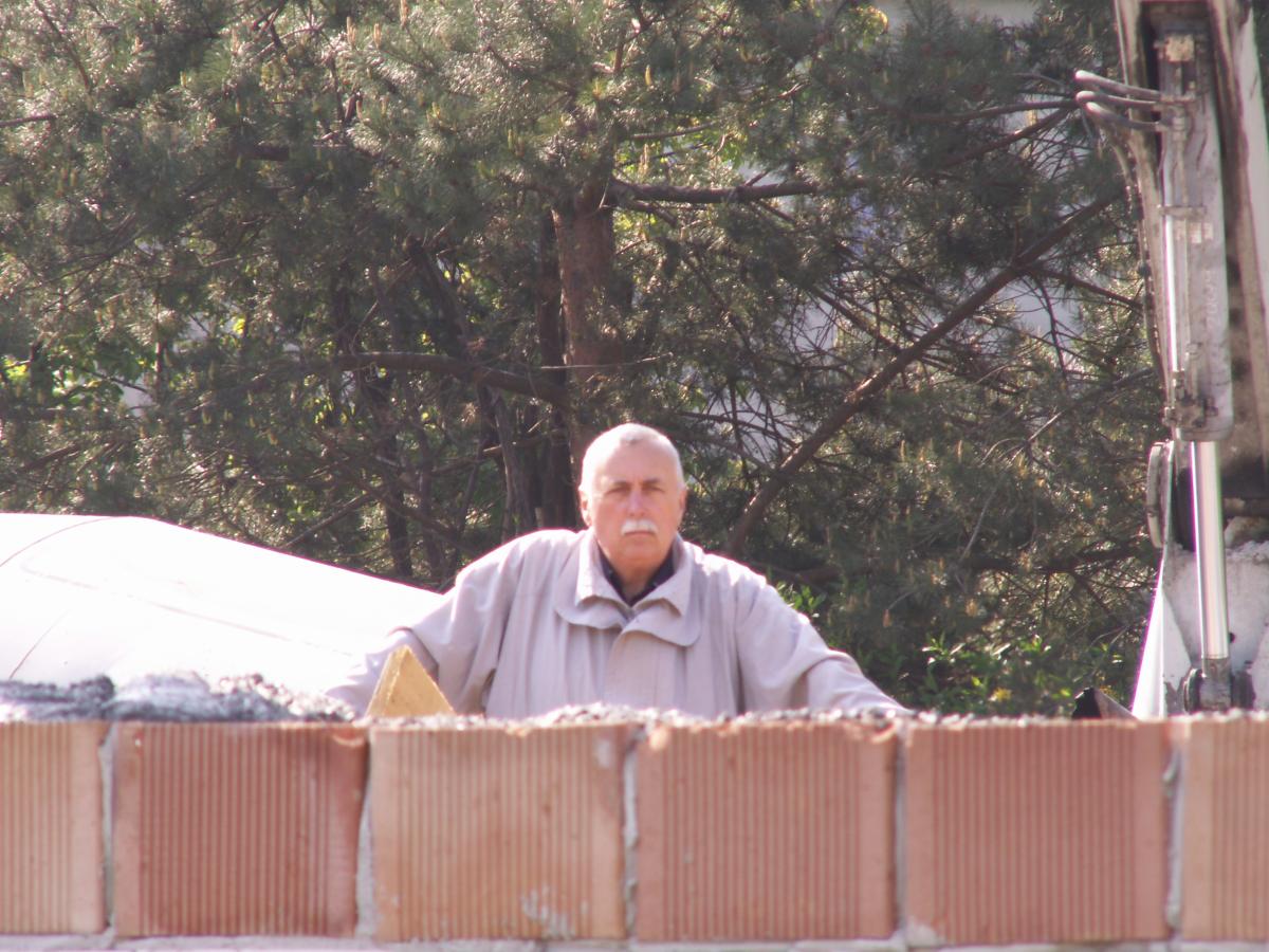 Mężczyzna w jasnej kurtce, z wąsami, stojący za murem z czerwonej cegły podczas budowy, w tle drzewa i fragment maszyny budowlanej.