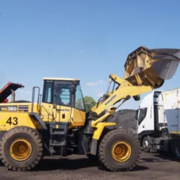 Żółta ładowarka Komatsu WA380 z pełną łyżką węgla, obok biała ciężarówka. W tle błękitne niebo i elementy sortowni. Praca na składzie opału.