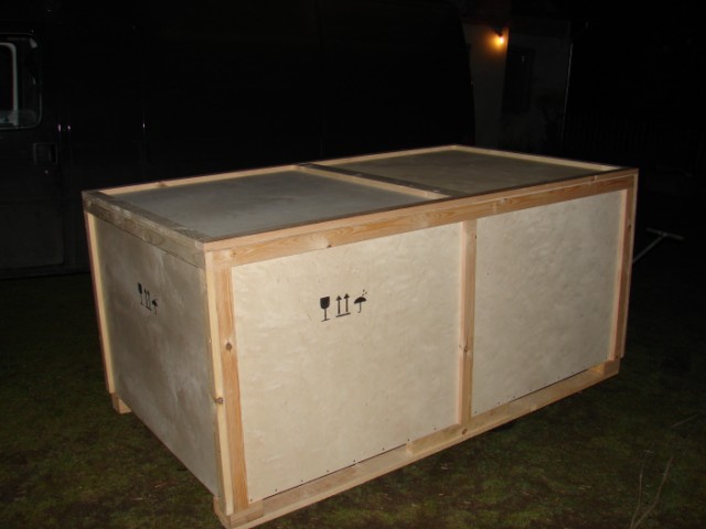 Duża, drewniana skrzynia transportowa na palecie, z oznaczeniami delikatnego ładunku, sfotografowana na zewnątrz w słabym oświetleniu nocnym, częściowo widoczny samochód dostawczy.