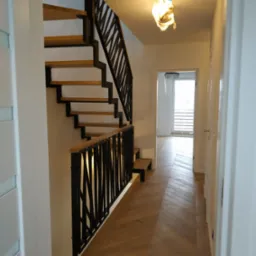 Nowoczesne schody drewniane z metalową balustradą w jasnym wnętrzu, parkiet ułożony we wzór jodełki, widok na korytarz z oknem.