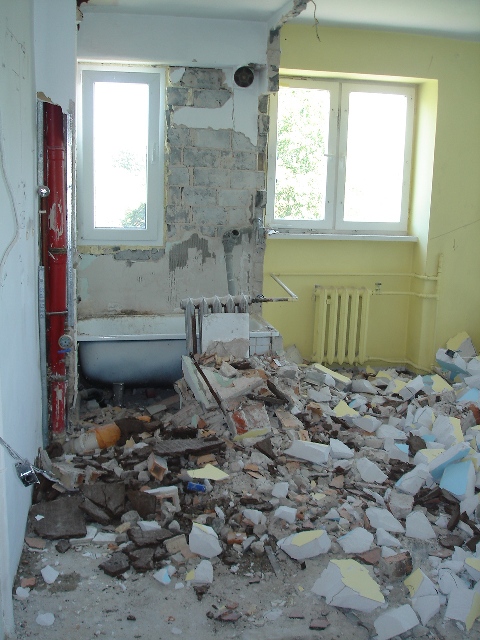 Łazienka w trakcie demontażu: gruz, odsłonięta cegła, fragment wanny i grzejniki. Widoczne rury instalacji wodnej. Dwa okna w tle.
