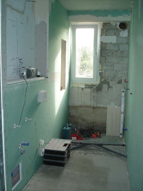 Remontowana łazienka w trakcie prac: ściany pokryte zielonymi płytami gipsowo-kartonowymi, widoczne instalacje wodno-kanalizacyjne, okno z widokiem na zieleń, narzędzia i materiały budowlane w tle,...