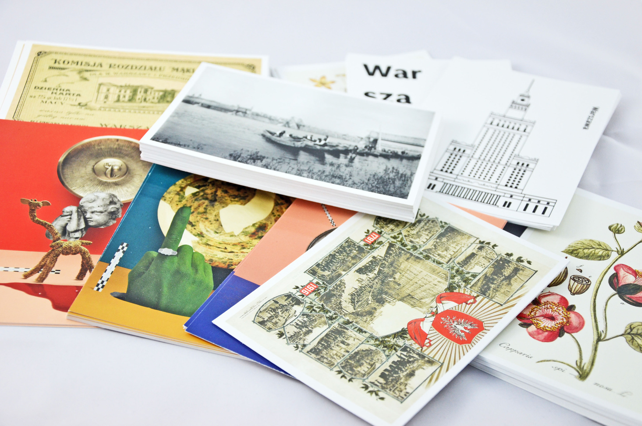 Stos kartek pocztowych i gadżetów reklamowych z motywami Warszawy, w tym zdjęcia czarno-białe, grafiki i ilustracje botaniczne na białym tle.