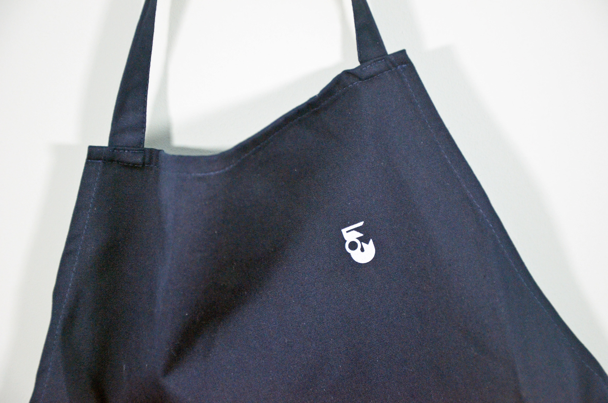 Granatowy fartuch z białym logo, zawieszony na wieszaku, ukazujący detale szycia i minimalistyczny design. Widoczne szwy i faktura materiału.