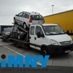 Laweta Iveco z trzema samochodami na platformie, zaparkowana obok żółtej naczepy. Widoczny fragment niebieskiej ciężarówki. Szare niebo.