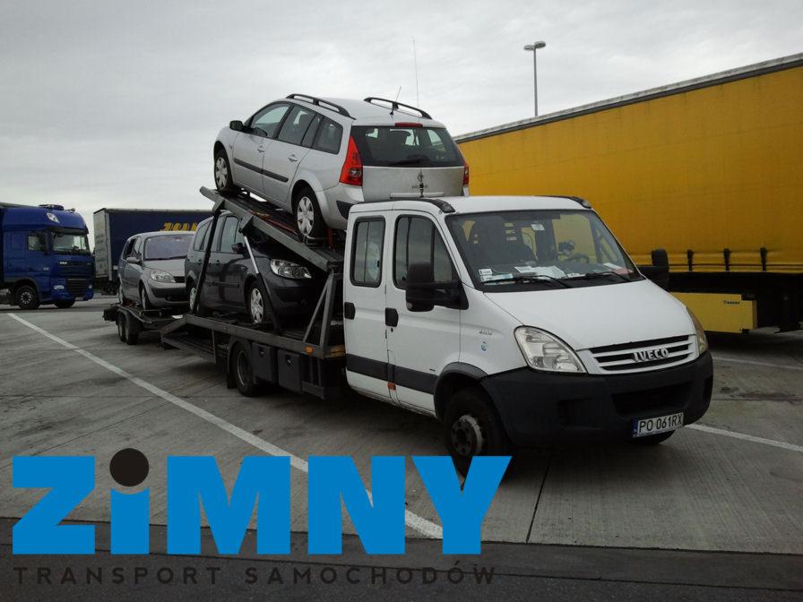 Laweta Iveco z trzema samochodami na platformie, zaparkowana obok żółtej naczepy. Widoczny fragment niebieskiej ciężarówki. Szare niebo.