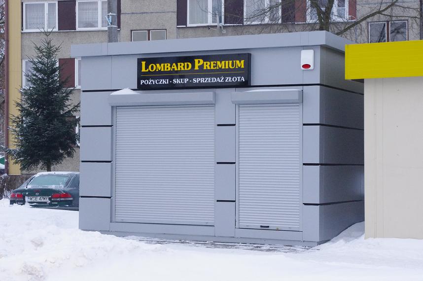 Szary pawilon handlowy z roletami antywłamaniowymi i szyldem 'Lombard Premium' na tle zimowego krajobrazu miejskiego.