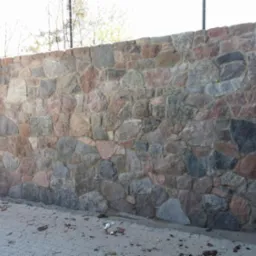 Murek oporowy z nieregularnych kamieni polnych w odcieniach szarości i czerwieni, z widocznym fragmentem chodnika z kostki brukowej u podstawy.