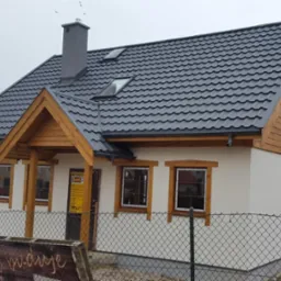 Nowoczesny dom szkieletowy z ciemnym dachem i drewnianymi elementami wykończeniowymi w Rokocinie. Widoczne okna dachowe i komin.