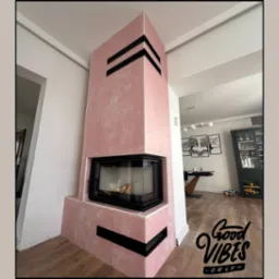 Różowy kominek z czarnymi akcentami w nowoczesnym wnętrzu. Widoczne drewno w palenisku i elementy dekoracyjne w tle. Nietypowe wykończenie kominka.