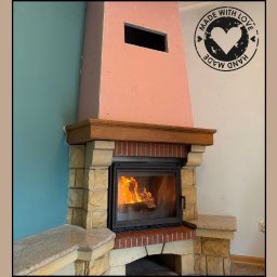 ART FIRE Nowicki kominki Sp. z o.o. - Otwarty kominek z paleniskiem, obudowany cegłą i kamieniem, z drewnianą półką. Widoczne płomienie i dekoracyjny napis 'Hand Made with Love'.