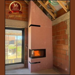ART FIRE Nowicki kominki Sp. z o.o. - Wnętrze w trakcie wykańczania z kominkiem obudowanym płytami gipsowymi. Widoczne palenisko z płomieniami, surowa cegła i okno z widokiem na zewnątrz.