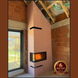 ART FIRE Nowicki kominki Sp. z o.o. - Narożny kominek obudowany płytami gipsowymi w surowym stanie, z widocznym paleniskiem i logo Art Fire, na tle ceglanej ściany i drewnianej konstrukcji dachu.