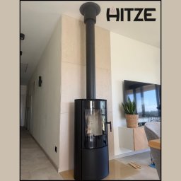 ART FIRE Nowicki kominki Sp. z o.o. - Stylowy, czarny piec wolnostojący z panoramiczną szybą, zintegrowany z czarnym przewodem kominowym, umieszczony na przezroczystej podkładce w nowoczesnym salonie z jasną ścianą dekoracyjną.