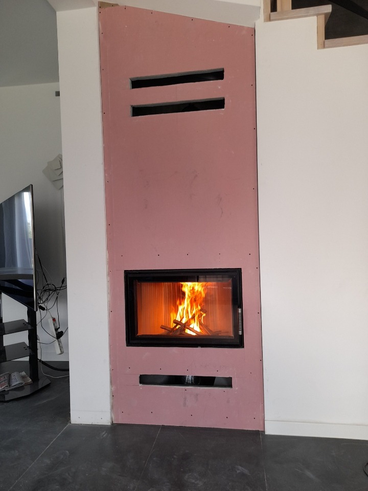 Wkład kominkowy z paleniskiem w trakcie montażu, obudowany płytami gipsowo-kartonowymi z widocznymi otworami wentylacyjnymi, na podłodze ciemne płytki.