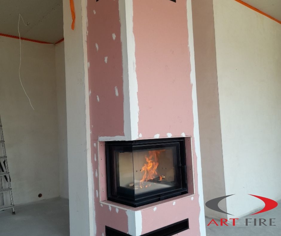 Niedokończona obudowa kominka z płyt gipsowo-kartonowych w kolorze różowym, widoczne białe spoiny, wkład kominkowy z palącym się ogniem, logo firmy w prawym dolnym rogu, drabina w tle.