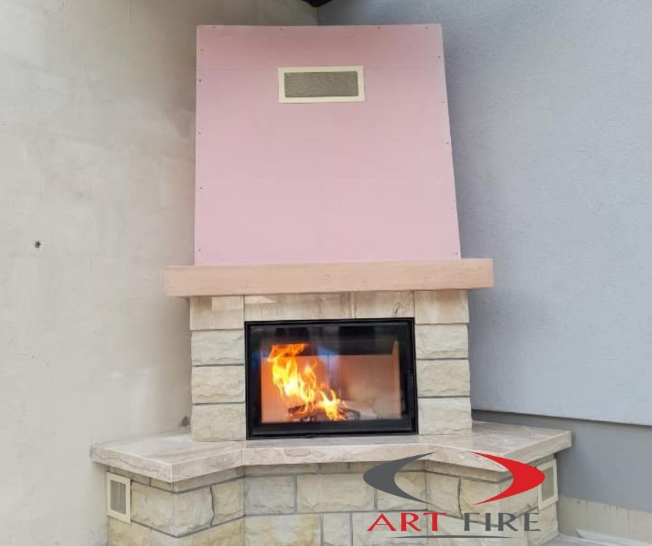 Narożny kominek z obudową z jasnego kamienia, czarną ramą paleniska i płomieniami wewnątrz, z fragmentem różowej płyty kartonowo-gipsowej powyżej, z logo Art Fire w prawym dolnym rogu.
