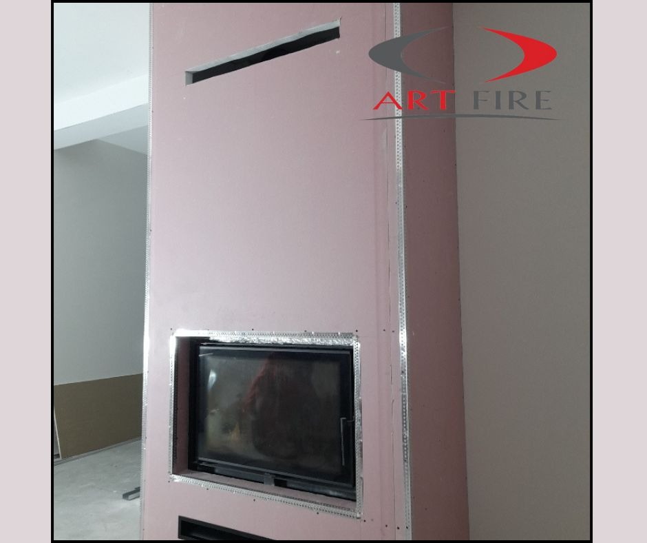 Wnętrze w trakcie wykończenia z widocznym wkładem kominkowym obudowanym płytami gipsowo-kartonowymi i logo firmy Art Fire w prawym górnym rogu.