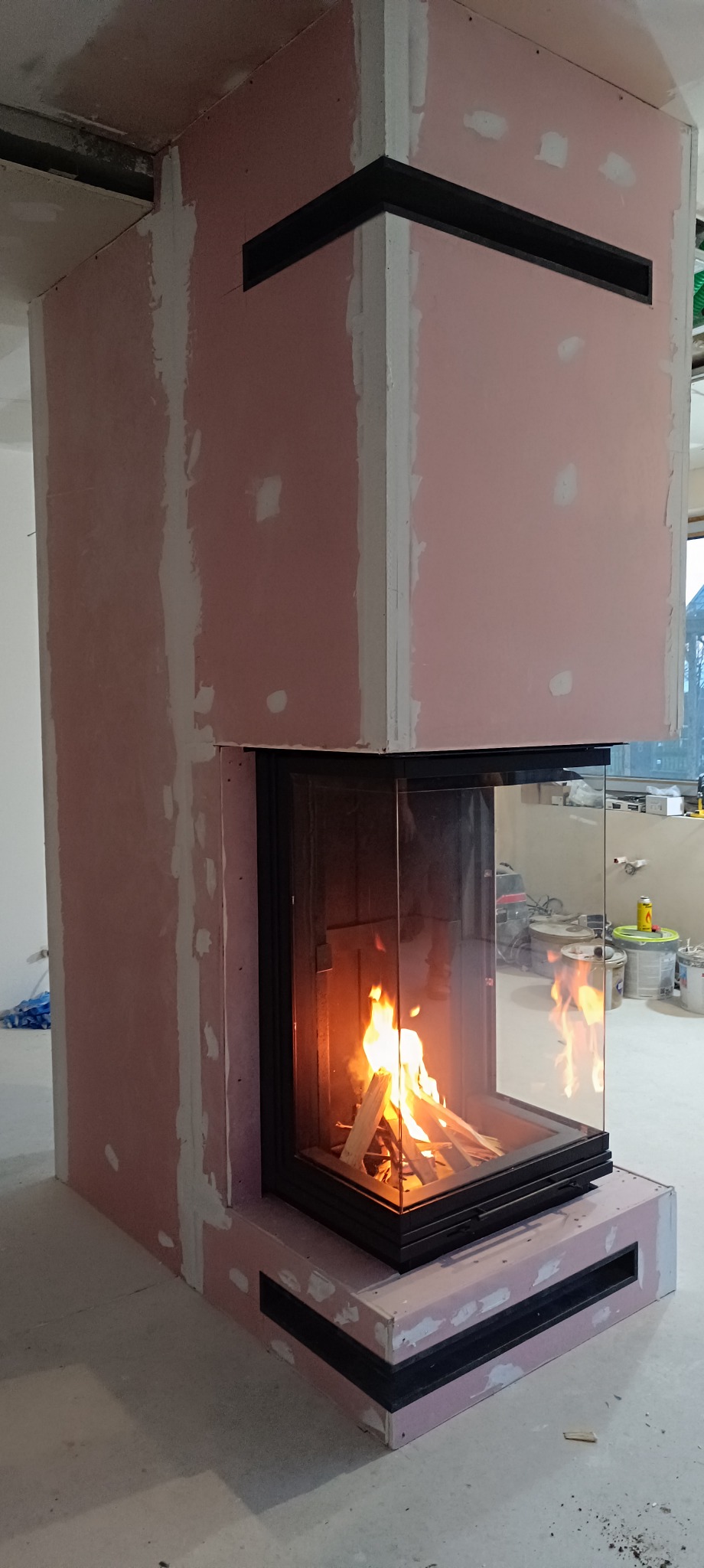Nowoczesny kominek narożny z szybą, w trakcie zabudowy płytami gipsowo-kartonowymi, widoczne szczeliny wentylacyjne w obudowie, płomienie w palenisku.