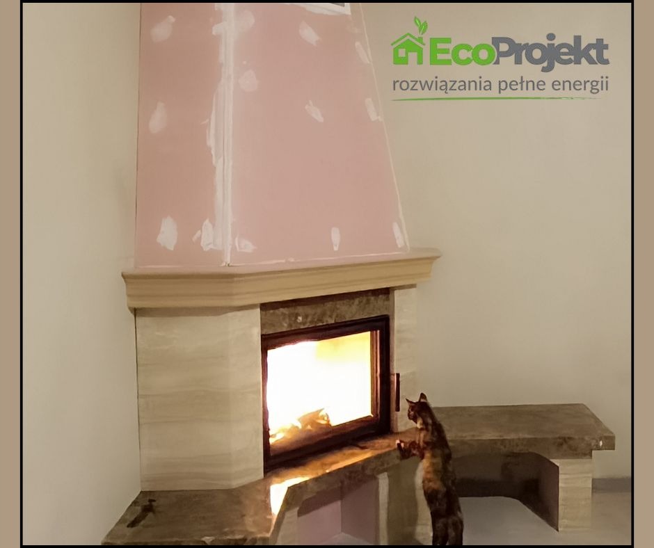 Kominek z marmurową obudową w trakcie wykańczania, z paleniskiem i siedzącym przed nim kotem. Widoczne logo firmy EcoProjekt.