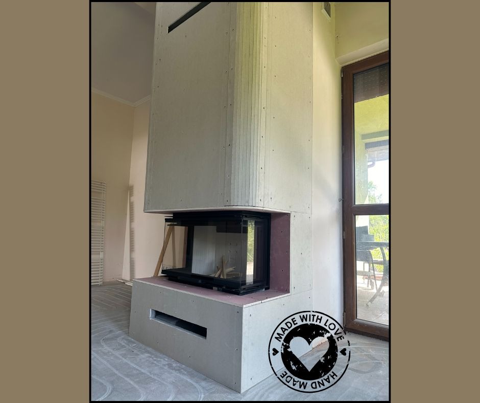 Wnętrze z kominkiem w obudowie z płyt gipsowych, widoczne instalacje ogrzewania podłogowego, okno z widokiem na zieleń. Etap wykończenia.