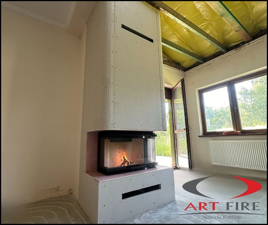 Wnętrze z kominkiem w trakcie obudowy płytami gipsowo-kartonowymi. Widoczne palenisko z płomieniami, instalacja ogrzewania podłogowego i surowy strop.