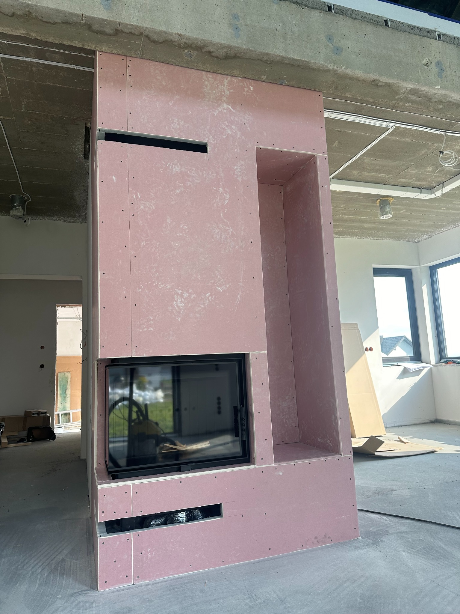 Konstrukcja obudowy kominka z płyt karton-gips, widoczne otwory wentylacyjne i wnęka, surowy betonowy sufit, w tle okno i elementy wykończeniowe.