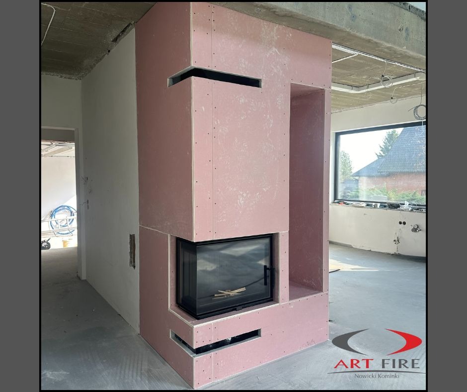 Surowy kominek obudowany płytami gipsowymi z czarnym wkładem, w trakcie montażu w jasnym wnętrzu z dużym oknem. Widoczne elementy konstrukcyjne.