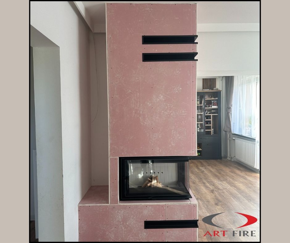 Wnętrze z kominkiem w trakcie obudowy płytami gipsowymi, widoczne czarne elementy dekoracyjne i palenisko. W tle okno i meble. Etap prac wykończeniowych.