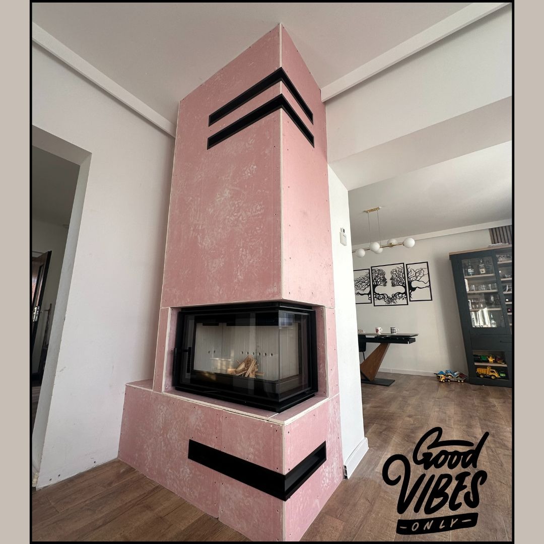 Różowy kominek z czarnymi akcentami w nowoczesnym wnętrzu. Widoczne drewno w palenisku i elementy dekoracyjne w tle. Nietypowe wykończenie kominka.