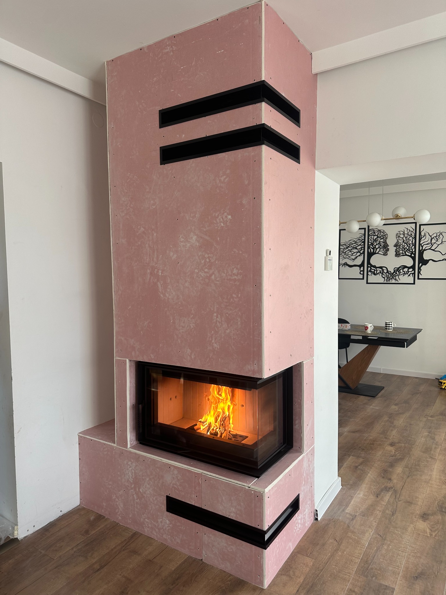 Wnętrze z kominkiem w trakcie obudowy z płyt gipsowych w kolorze różowym, z widocznym paleniskiem i czarnymi elementami dekoracyjnymi. W tle jadalnia z nowoczesnym oświetleniem.