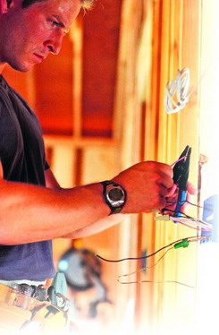 Elektryk w trakcie pracy, używa szczypiec do przewodów w instalacji elektrycznej w nowo budowanym domu, widoczne drewniane elementy konstrukcyjne, pasek z narzędziami i zegarek na ręce.