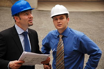 Dwóch mężczyzn w kaskach, jeden w garniturze trzyma plan instalacji elektrycznej, drugi w koszuli patrzy w obiektyw, tło stanowi brukowana powierzchnia.