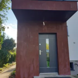 Wejście do domu z nowoczesnymi drzwiami w kolorze grafitowym, elewacja w odcieniach czerwieni i brązu, widoczna niedokończona instalacja elektryczna oświetlenia zewnętrznego.