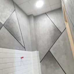 Ściana z dekoracyjnym tynkiem w geometryczne wzory, oddzielona białymi kafelkami. Widoczny fragment sufitu i drewniany element wykończeniowy.