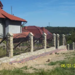 Nowoczesne ogrodzenie z kutymi elementami i kamiennymi słupkami, świeżo po montażu. Widoczny fragment domu z czerwoną dachówką i ścieżka z kostki.