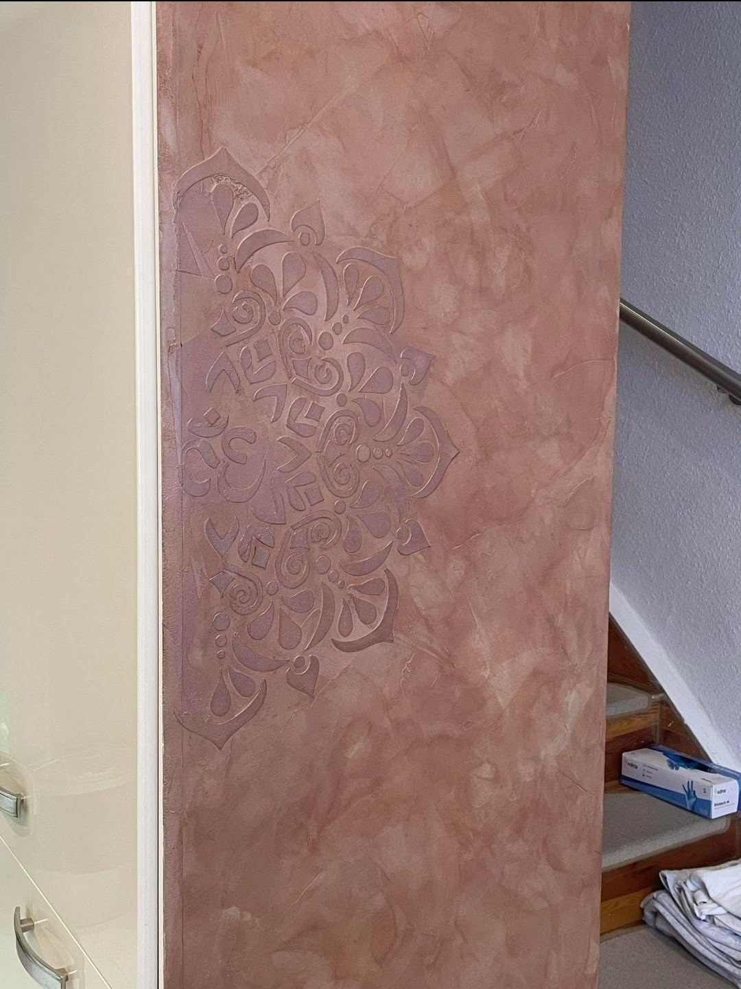 Różowa ściana z dekoracyjnym tynkiem strukturalnym w stylu mandali, widoczna częściowo szafka i schody w tle. Efektowna dekoracja ściany wewnątrz budynku.