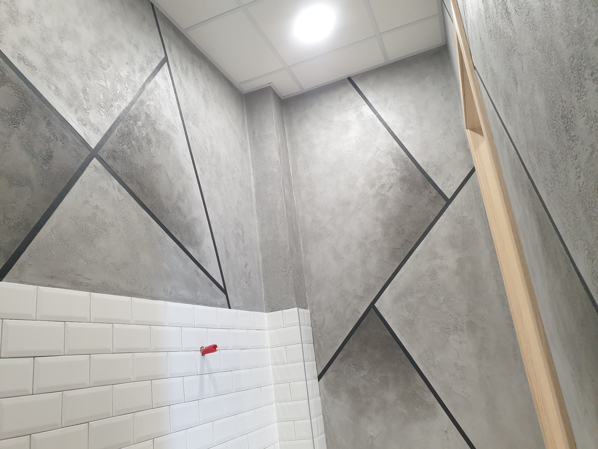 Ściana z dekoracyjnym tynkiem w geometryczne wzory, oddzielona białymi kafelkami. Widoczny fragment sufitu i drewniany element wykończeniowy.